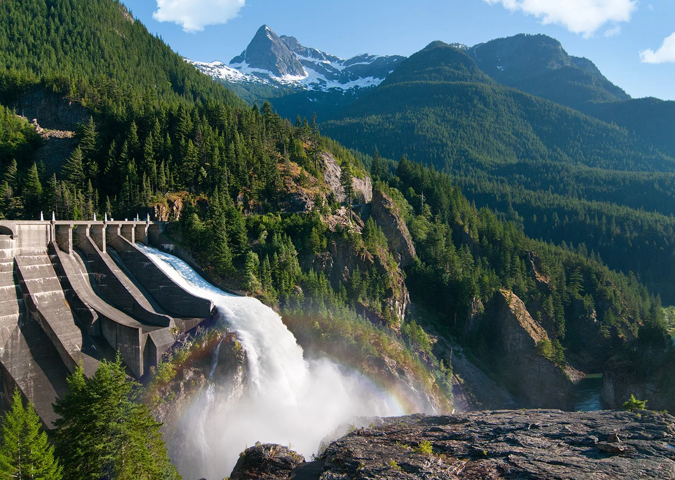  Ein Wasserkraftwerk in alpiner Umgebung mit staubedingtem Wasserabfluss, Sprühnebel und Regenbogen vor bewaldeter Gebirgskulisse.