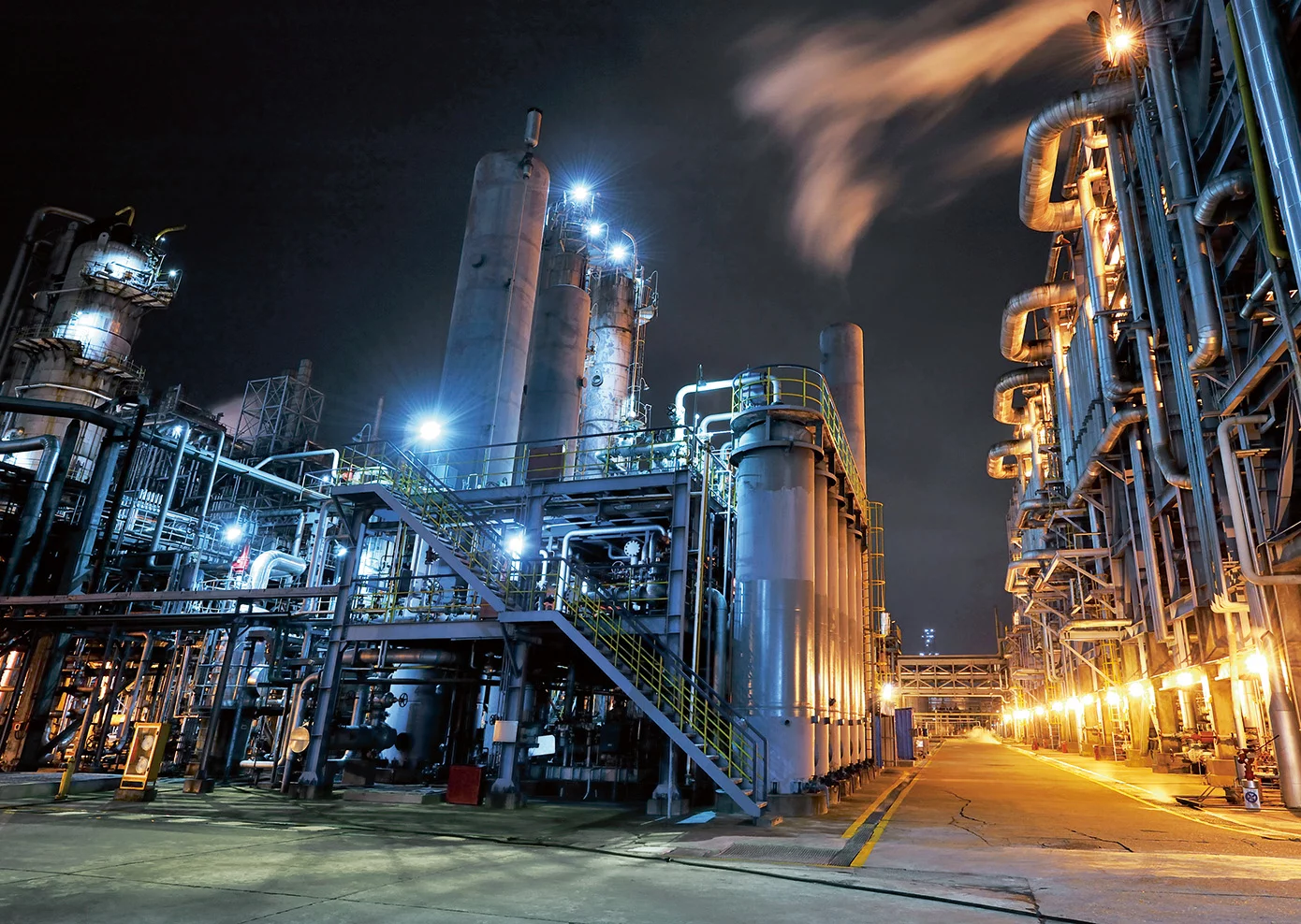  Beleuchtete Industrieanlage mit Rohrleitungen, Türmen und Dampfaustritt bei Nacht – vermutlich aus der Chemie-, Petrochemie- oder Energiebranche.