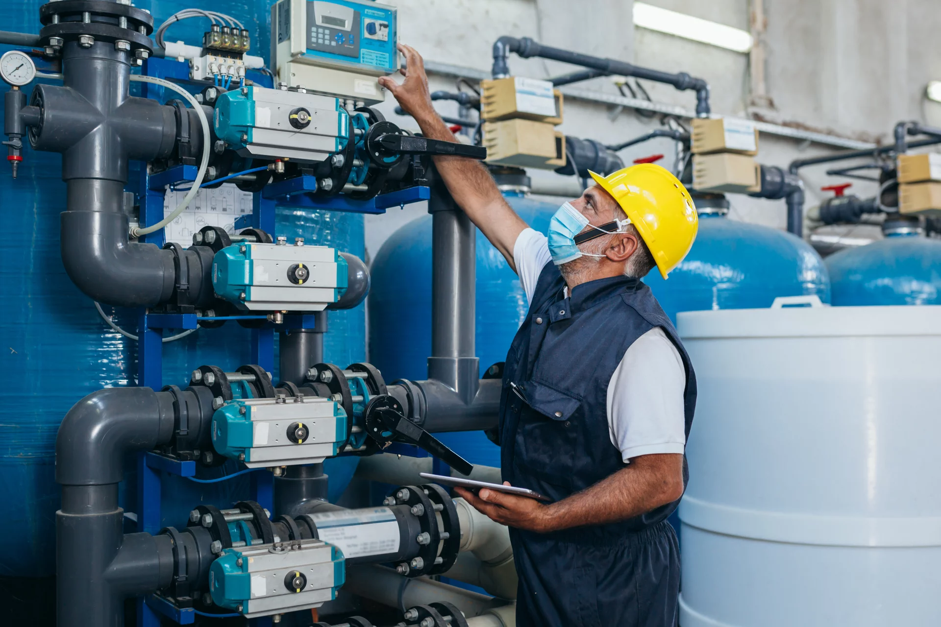  Ein Mitarbeiter mit Schutzmaske und Helm überprüft eine Wasseraufbereitungsanlage über ein digitales Steuergerät.
