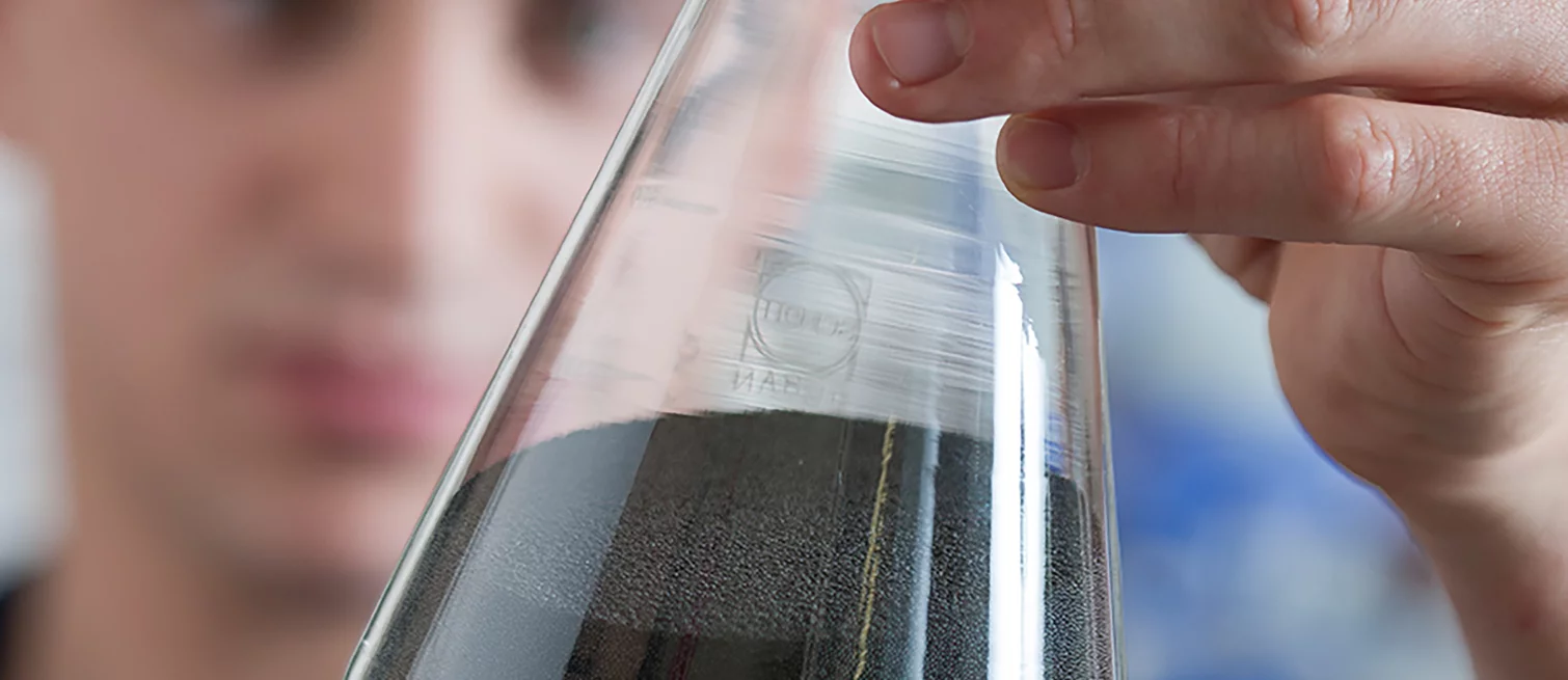  An employee examines a laboratory Erlenmeyer flask containing fine-grained black material, likely graphite or powder.