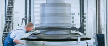  Factory worker inspecting a large cylindrical component made of graphite or technical ceramic on an industrial machine.