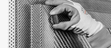 Ein Mitarbeiter mit Handschuhen prüft die Oberfläche eines Bauteils aus carbonfaserverstärktem Kohlenstoff mit typischer Gewebestruktur.