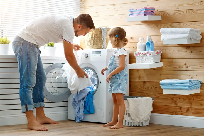  Father and daughter load a washing machine in a bright, cosy laundry room with wooden panelling and neatly stacked textiles.