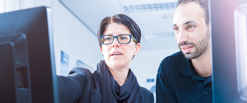  woman standing next to a man at a desk poiting at a screen