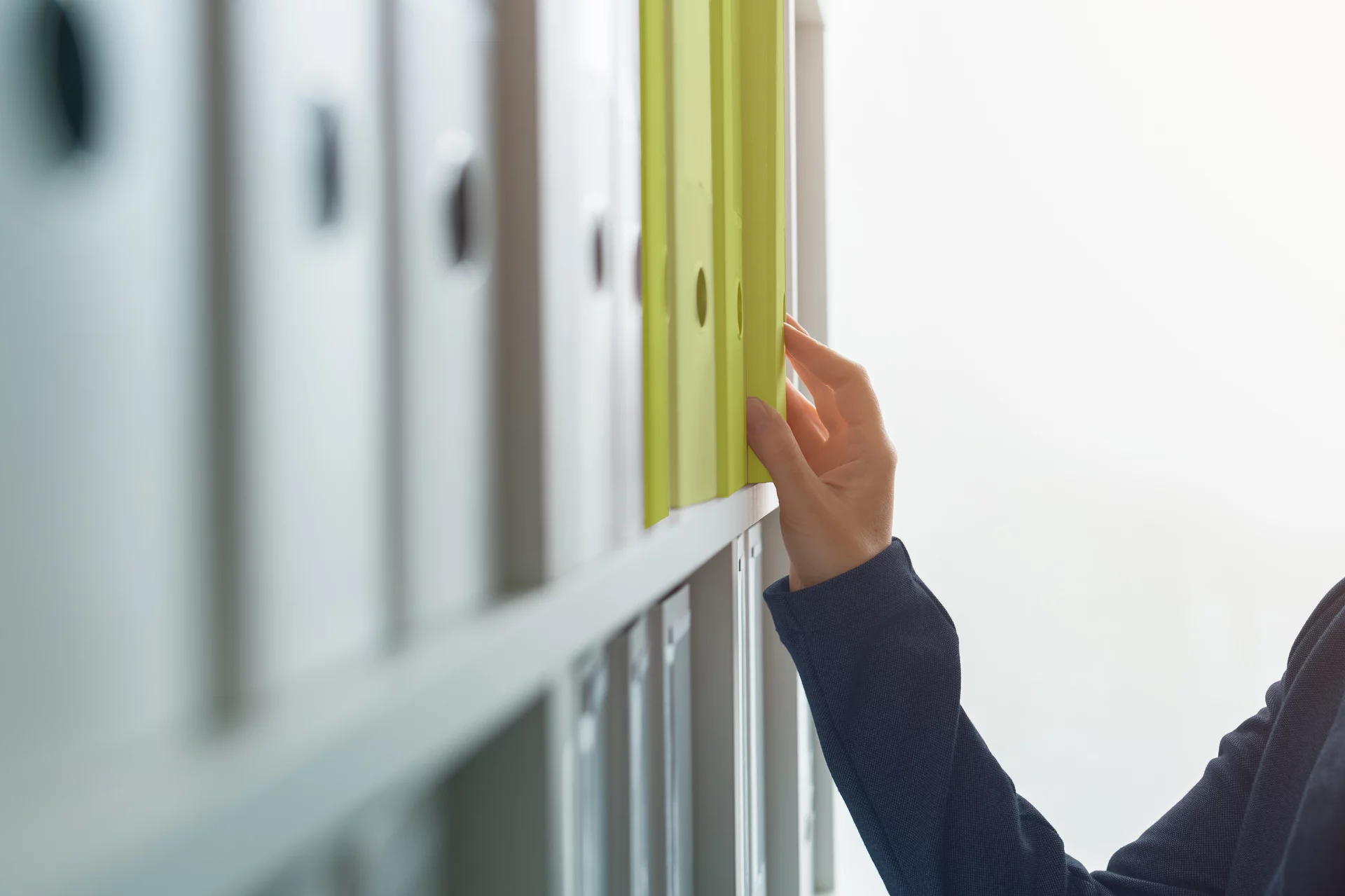 Eine Person greift in ein Regal im Büro mit diversen Ordnern und greift sich einen bestimmten Ordner
