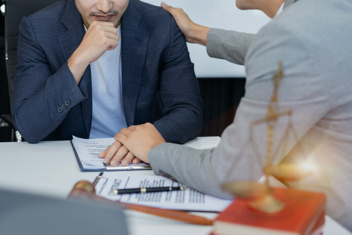  Two people are sitting at a table. One person puts a consoling hand on the other's shoulder.