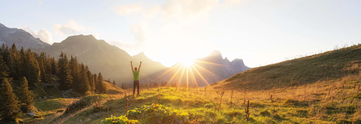  A person stands in the green on a hill and watches the sunrise in the alpine panorama