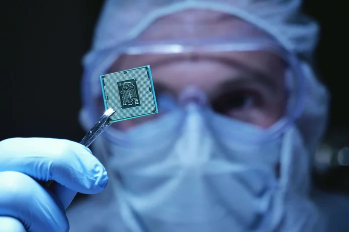  Person in protective clothing is looking at a microchip, which he is holding with tweezers.