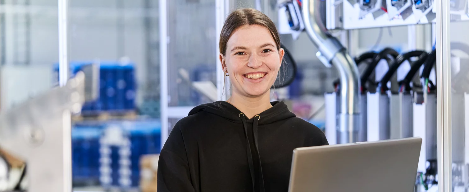 Studierende im Bereich System Engineering mit Laptop in den Händen arbeitet an einer Maschine